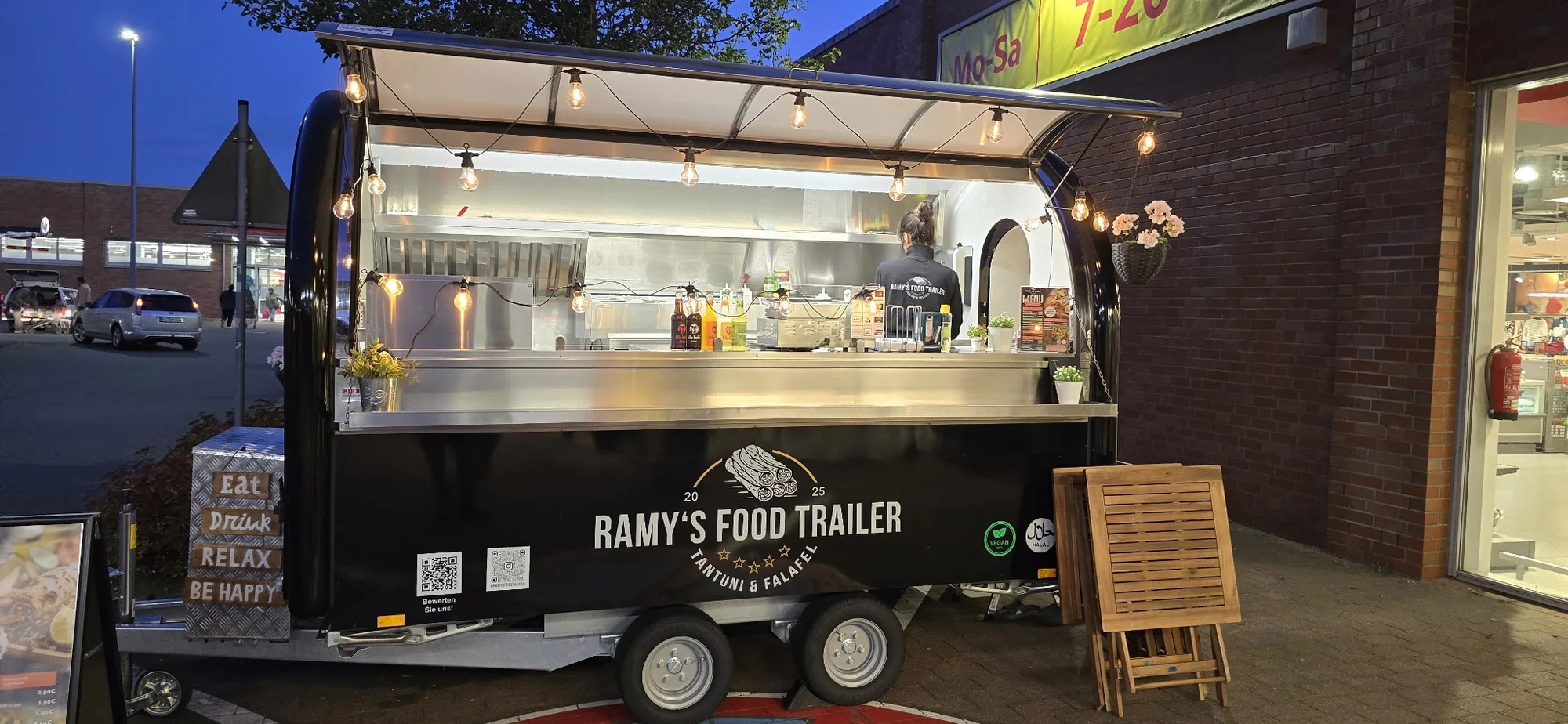 Ramy's Food Trailer am Abend, die beleuchtete Ausgabetheke und eine Lichterkette über dem Wagen