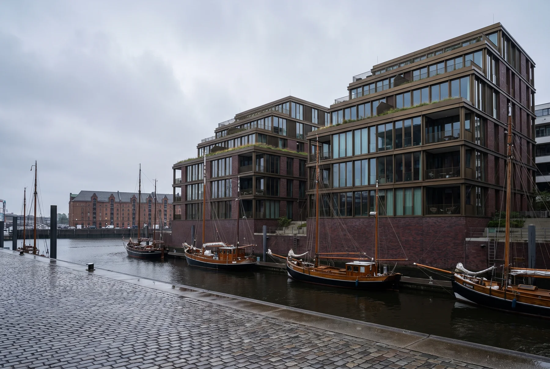 Backsteinbau mit bronzefarbenen Fenstern am Hafenbecken, davor liegen Traditionssegler