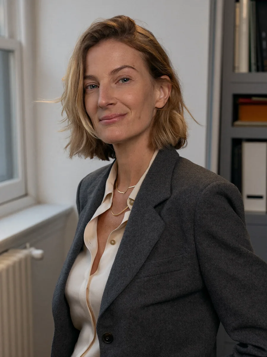 Frau in dunkelgrauem Blazer und cremefarbener Bluse am Fenster eines Büros